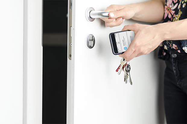 One hand is opening a door while the other is holding a smartphone with the Klevio app and a bunch of physical keys