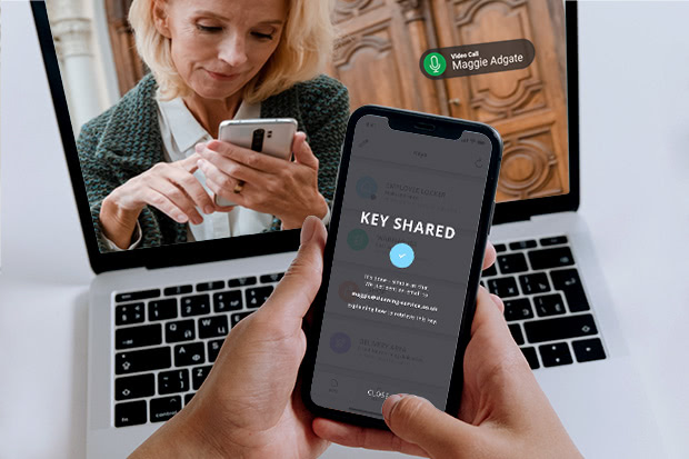 A hand holding a phone with a smart lock app that shows a key has been shared while the recipient is visible on a video call in the background