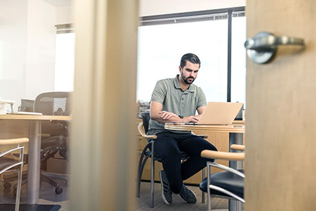 A view from a slightly opene door of an office worker sitting behind a desk and working on his laptop