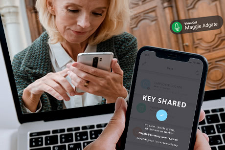 Detail of a smartphone with a smart lock app showing that keys have been successfully shared while the key receiver is shown in the background on a video call at some other location, accepting these keys on their own smartphone