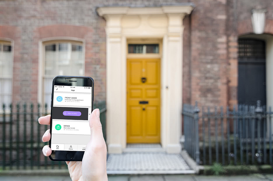 Hand holding up a smart lock app and a house with a yellow door in the background